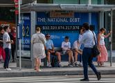 Pedestrians sit at a bus shelter at Pennsylvania Avenue and 22nd Street NW where an electronic billboard and a poster display the current U.S. National debt per person and as a nation at 32 Trillion dollars on July 05, 2023 in Washington, DC.